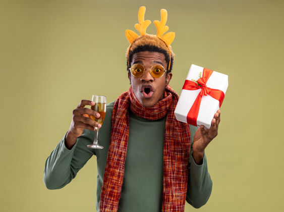 礼物一个非洲裔美国人 戴着滑稽的边 戴着鹿角 脖子上围着围巾 手里拿着一杯香槟和圣诞礼物 站在绿色的背景下惊讶地看着摄像机围巾香槟角