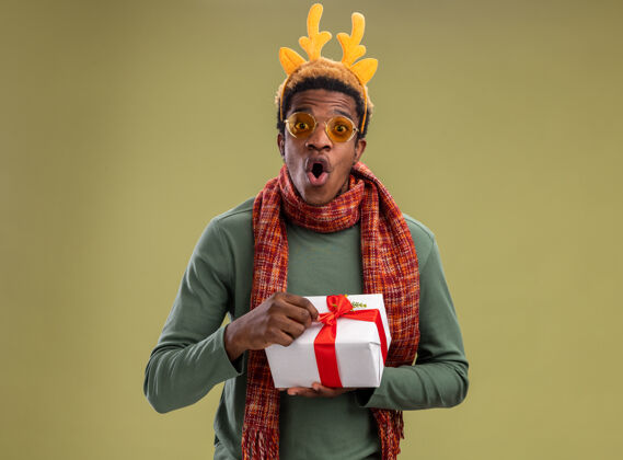 鹿一个非洲裔美国人 戴着滑稽的边 戴着鹿角 脖子上围着围巾 手里拿着圣诞礼物 站在绿色的背景上惊讶地看着摄像机非洲脖子圣诞节