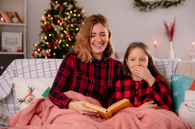 女儿笑眯眯的母女俩坐在沙发上读着铺着毛毯的书 在家里享受圣诞节时光享受妈妈封面