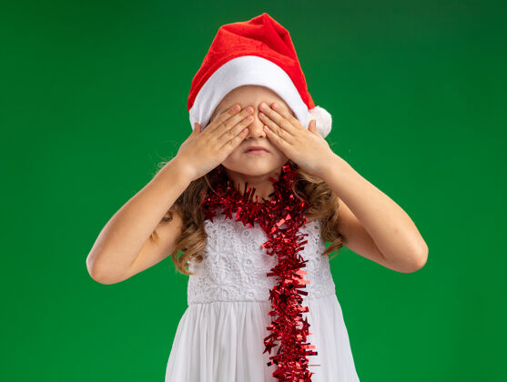 小小女孩戴着圣诞帽 脖子上戴着花环 眼睛蒙着 手被隔离在绿色的墙上戴圣诞节盖
