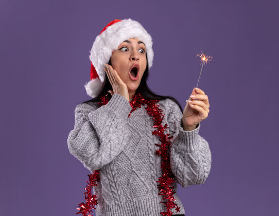 金属箔令人印象深刻的年轻白人女孩戴着圣诞帽 脖子上戴着金箔花环 手放在脸上 手放在紫色的墙上 留着复制空间脖子帽子穿着