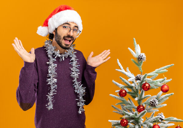 戴着快乐的年轻帅哥站在圣诞树旁 戴着圣诞帽 脖子上戴着花环 双手分开放在橙色的墙上圣诞帅气花环