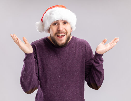 年轻穿着紫色毛衣 戴着圣诞帽的年轻人看着摄像机 困惑地微笑着 双臂伸向两边 站在白色的背景上毛衣圣诞老人伸展