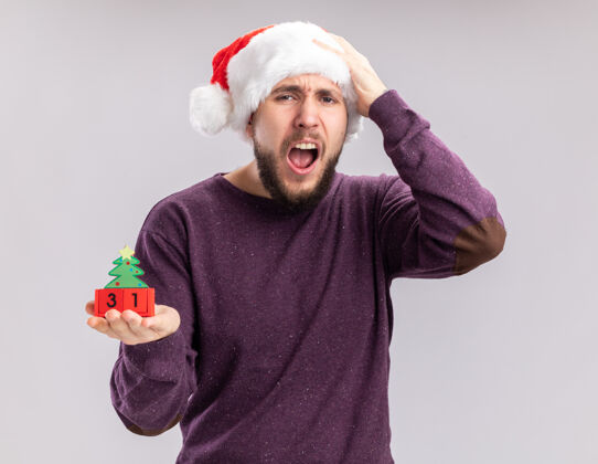 年穿着紫色毛衣 戴着圣诞帽的年轻人站在白色背景上 带着失望的表情高喊着新年约会的方块立方体圣诞老人站着