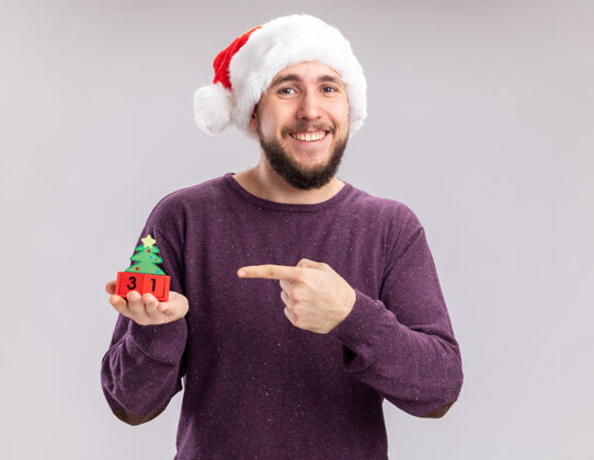 帽子身穿紫色毛衣 头戴圣诞帽的年轻人站在白色背景上 用食指指着方块上的新年日期 快乐而愉快圣诞老人立方体手指