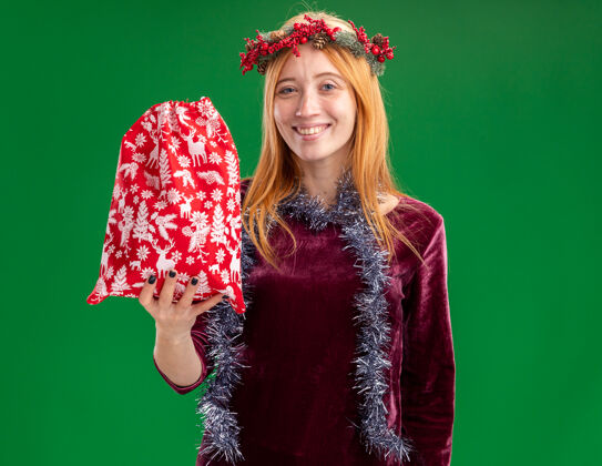 包微笑着的年轻漂亮的女孩穿着红色的裙子 脖子上戴着花环和花环 手里拿着绿色背景上孤立的圣诞包圣诞节抱着脖子