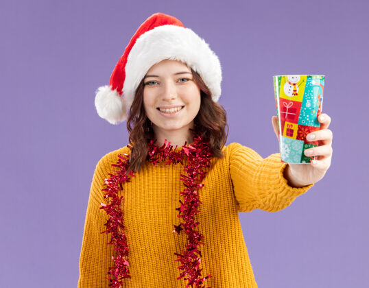 杯子微笑着的年轻斯拉夫女孩 戴着圣诞帽 脖子上戴着花环 手里拿着纸杯 背景是紫色 有复印空间脖子花环帽子