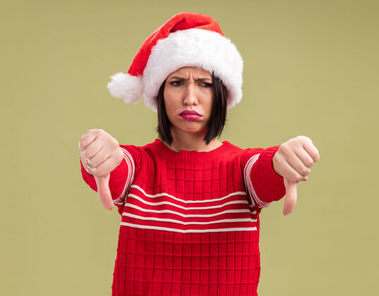 向下愁眉苦脸的年轻女孩戴着圣诞帽 看着一边竖起大拇指 孤立地站在橄榄绿的背景上拇指侧圣诞老人