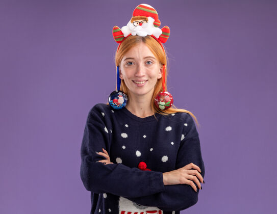 耳朵微笑着的年轻漂亮女孩穿着圣诞毛衣 戴着圣诞发箍 耳朵上挂着圣诞球 双手交叉 孤立在紫色背景上交叉手女孩