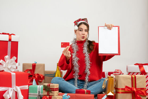 文件带着圣诞帽的派对女孩拿着卡片和文件坐在白色的礼物旁边购物肖像坐