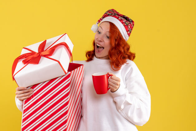 人黄色墙上年轻女子手持圣诞礼物和一杯茶的正面图女性礼物快乐