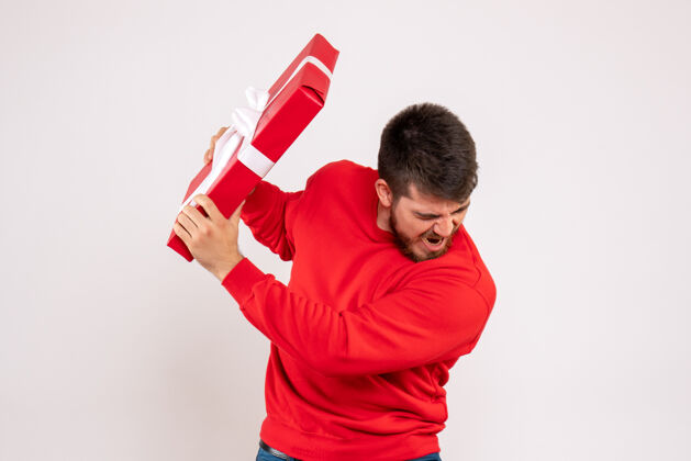节日一个穿着红衫的年轻人拿着圣诞礼物站在白色的墙上手微笑微笑
