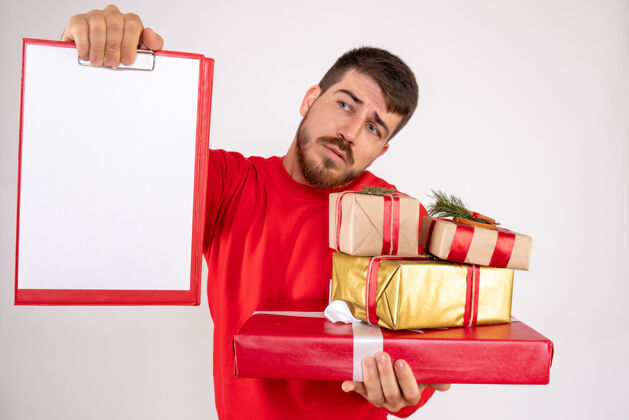文件身穿红衫的年轻人拿着圣诞礼物和白墙上的文件说明的正面视图微笑年轻男性年轻