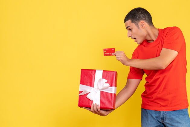 成人年轻人拿着礼物和银行卡在黄色墙上的正面图十二月圣诞节人