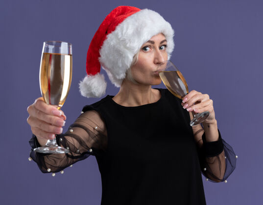 快乐戴着圣诞帽的中年金发女人看着相机 手里拿着两杯香槟 一边喝着一杯 一边伸开另一杯 对着紫色背景上孤立的相机朝着另一个金发