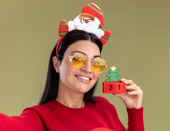 穿着特写镜头：微笑的白人女孩戴着圣诞老人的头带和带眼镜的毛衣 手里拿着圣诞树玩具 橄榄绿的墙上挂着被隔离的日期圣诞节橄榄毛衣
