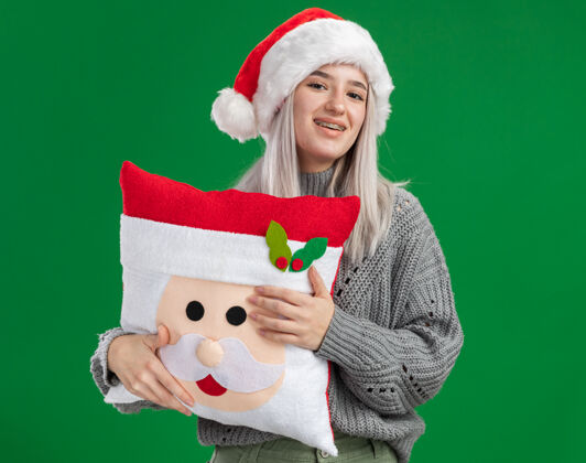 抱着快乐的金发女郎 穿着冬衣 戴着圣诞帽 抱着圣诞枕头 面带微笑地看着相机 站在绿色的背景下毛衣脸微笑