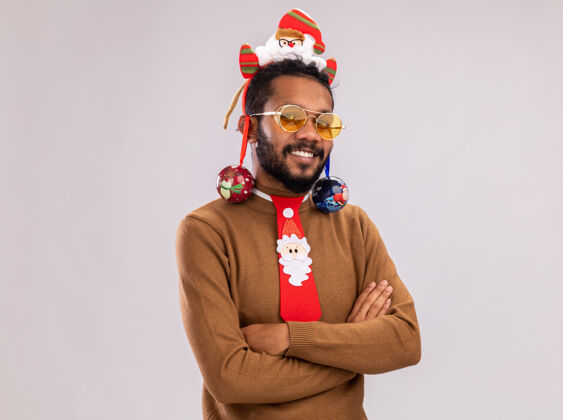 圣诞节快乐的非洲裔美国人身穿棕色毛衣 头戴圣诞老人戒指 打着有趣的红色领带 手里拿着圣诞球 站在白色背景下 面带微笑地看着摄像机脸球微笑