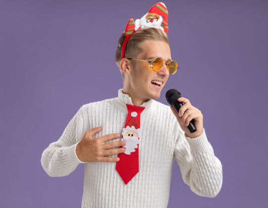 穿着快乐的年轻帅哥戴着圣诞老人的头带和领带戴着眼镜拿着麦克风手放在胸前看着边唱隔离在紫色墙上年轻领带侧边