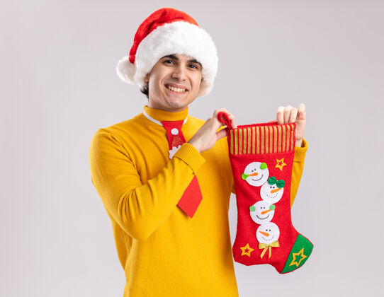 年轻快乐的年轻人穿着黄色高领毛衣 戴着圣诞老人帽 打着滑稽的领带 站在白色的背景上 露出欢快的笑容高领毛衣长袜圣诞节