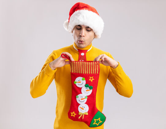 长袜穿着黄色高领毛衣 戴着圣诞老人帽 打着滑稽领带的年轻人拿着圣诞长袜 站在白色背景上看里面圣诞节里面领带