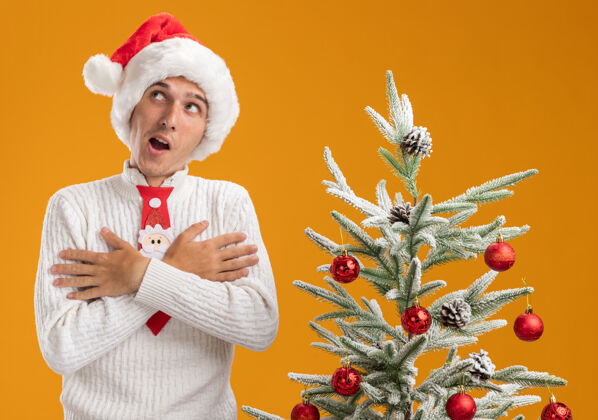站着令人印象深刻的年轻帅哥戴着圣诞帽 打着圣诞老人的领带 站在装饰好的圣诞树旁 双手交叉放在胸前 看着橙色墙上孤立的一面圣诞老人年轻帅气