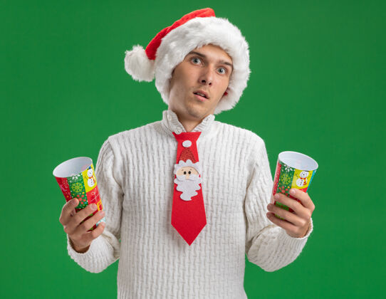 领带给人印象深刻的年轻帅哥戴着圣诞帽打着圣诞老人的领带拿着塑料圣诞杯隔离在绿色的墙上帽子圣诞老人年轻