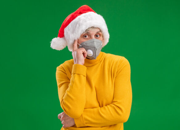 长相体贴的老妇人戴着医用面具戴着圣诞帽 把手指放在太阳穴上 看着隔离在绿色背景上的相机 还有复印空间手指体贴面具