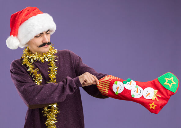 金属丝一个留着胡子的年轻人戴着圣诞老人的帽子 脖子上戴着金属丝 手里拿着圣诞长袜 站在紫色的背景下 看上去很困惑长袜围着脖子