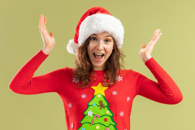 圣诞节穿着红色圣诞毛衣 戴着圣诞帽的年轻女子高兴而兴奋地举起双臂站在绿色的背景上毛衣站着圣诞老人