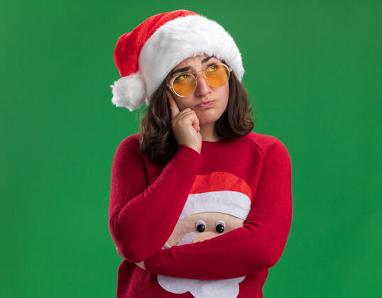 毛衣穿着圣诞毛衣 戴着圣诞帽和眼镜的年轻女孩站在绿色的墙上 困惑地抬头望去困惑圣诞老人年轻人