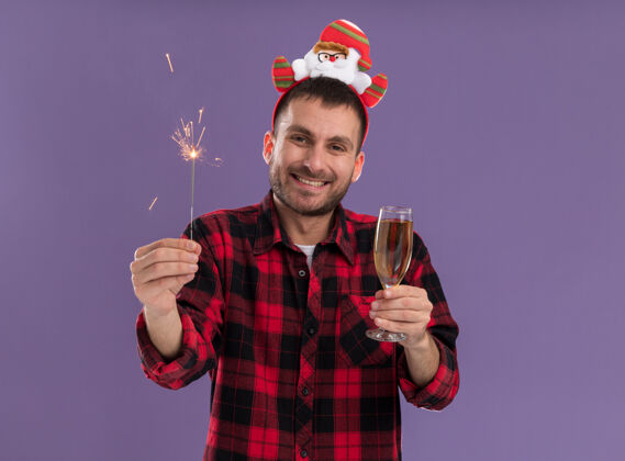 圣诞节快乐的白人年轻人戴着圣诞老人的头带 手里拿着节日火花和香槟 微笑着孤立地站在紫色的墙上 留着复制空间头带穿着玻璃杯