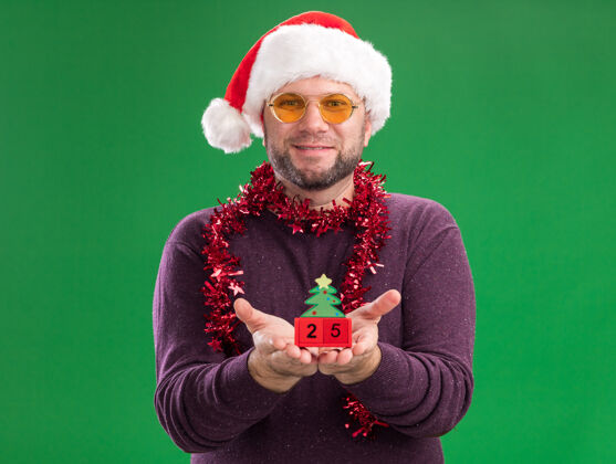 脖子高兴的中年男子戴着圣诞帽 脖子上戴着金箔花环 戴着眼镜 伸出圣诞树玩具 绿色的墙上有日期隔离圣诞节男人花环