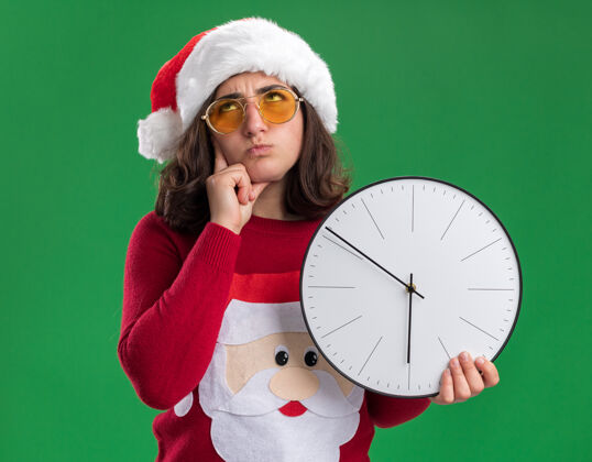 帽子穿着圣诞毛衣 戴着圣诞帽 戴着圣诞眼镜的小女孩举着挂钟 站在绿色的背景上 困惑地抬头看困惑年轻人圣诞节