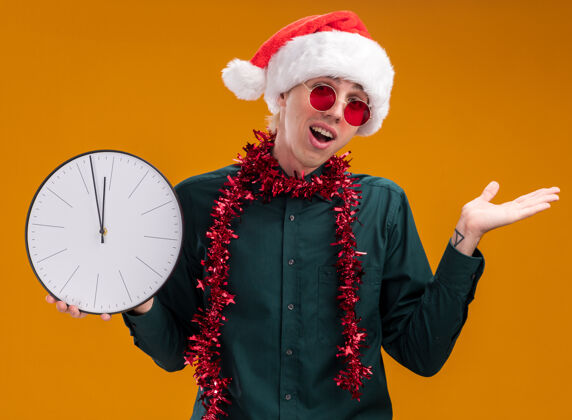 手令人印象深刻的年轻金发男子戴着圣诞帽 戴着眼镜 脖子上戴着金属丝花环 拿着时钟看着摄像机 镜头上显示的是孤立在橙色背景上的空手金发帽子周围