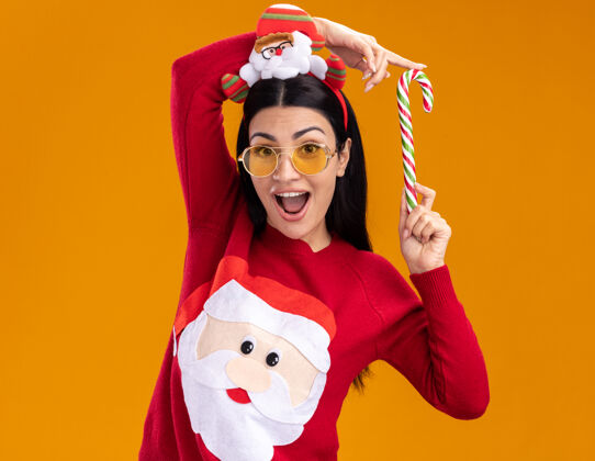 穿着令人印象深刻的年轻白人女孩戴着圣诞老人的头带和毛衣 戴着眼镜 手持传统的圣诞糖果手杖 靠近孤立在橙色墙上的头传统圣诞老人圣诞老人