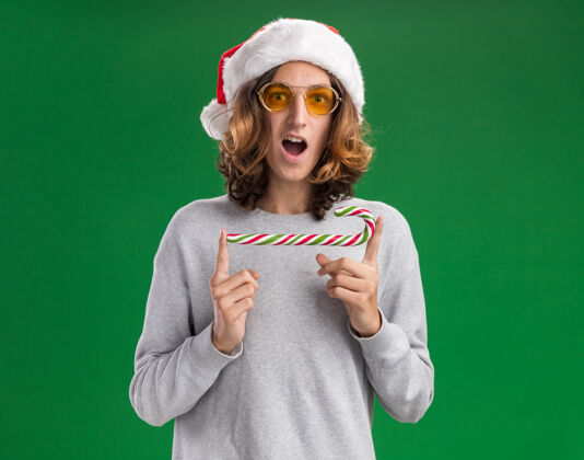 手杖快乐的年轻人戴着圣诞老人的帽子 戴着黄色的眼镜 拿着糖果手杖 站在绿色的背景上惊讶地看着镜头惊讶糖果惊讶