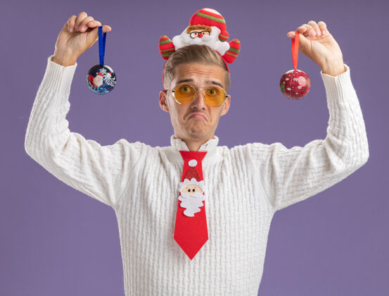 小伙子年轻帅哥戴着圣诞老人的头带和领带 戴着眼镜 举着圣诞球饰物 看着紫色背景上孤立的相机圣诞老人相机年轻人