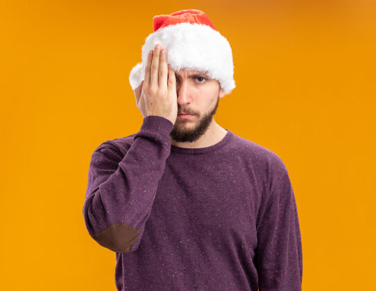 帽子一个穿着紫色毛衣 戴着圣诞老人帽子的年轻人 一只手站在橙色的背景上 遮住了一只眼睛站着年轻圣诞老人