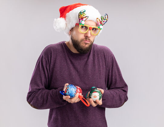 眼镜一个穿着紫色毛衣 戴着圣诞帽 戴着滑稽眼镜 拿着圣诞球的年轻人站在白色背景下 困惑而不确定地看着摄像机困惑毛衣圣诞节