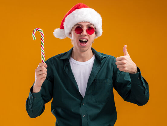 圣诞节令人印象深刻的年轻金发男子戴着圣诞帽 戴着眼镜 手持圣诞甜手杖 竖起大拇指 看着孤立在橙色背景上的相机圣诞老人拇指年轻