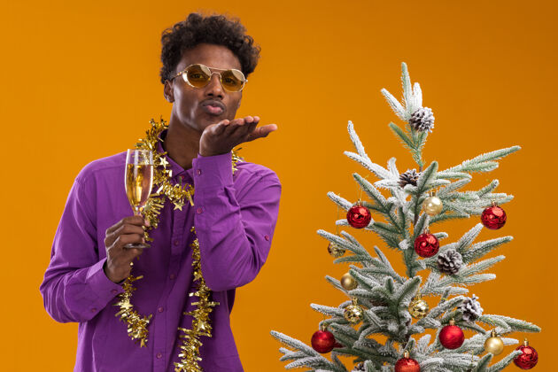 快乐自信的美国黑人年轻人戴着一副戴着金箔花环的眼镜 站在装饰好的圣诞树旁 手里拿着一杯香槟装饰非裔美国人圣诞快乐