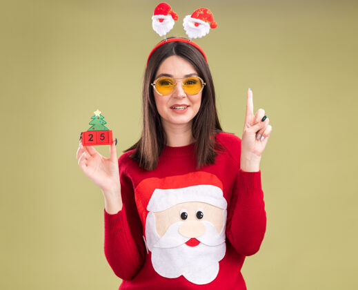 毛衣印象深刻的年轻漂亮的白人女孩穿着圣诞老人毛衣和头带眼镜举行圣诞树玩具与日期头带女孩圣诞老人