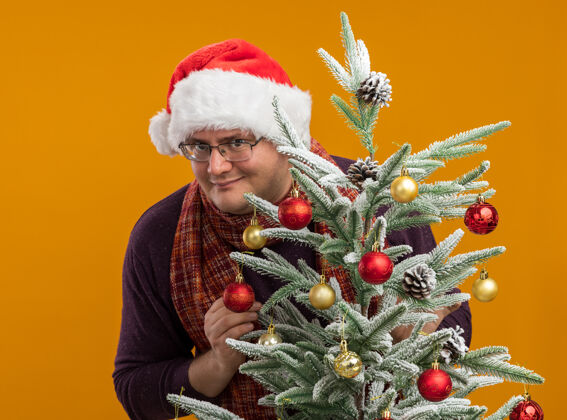 围巾戴着眼镜 戴着圣诞帽 脖子上围着围巾 站在装饰好的圣诞树后面 看着橙色背景上的相机 他很高兴圣诞树橙色围着