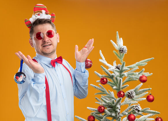 眼镜戴着吊带领结的年轻人站在圣诞树旁边 戴着圣诞老人和红眼镜 看起来很困惑 拿着玩具在橙色的背景下为圣诞树领带吊带圣诞节