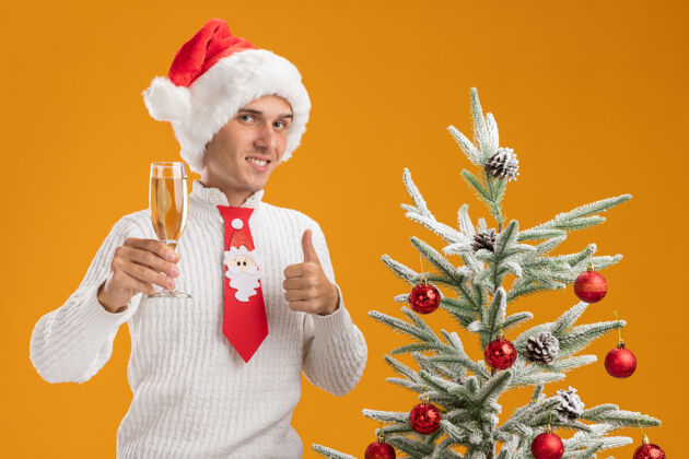 站着微笑着的年轻帅哥戴着圣诞帽 打着圣诞老人的领带 站在装饰好的圣诞树旁 手里拿着一杯香槟 看着相机 在橙色背景上孤立地竖起大拇指帽子玻璃帅气