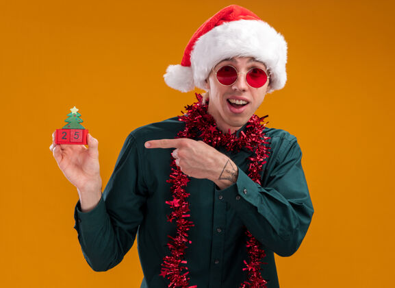 举行令人印象深刻的年轻金发男子戴着圣诞帽 戴着眼镜 脖子上戴着金属丝花环 拿着并指着圣诞树玩具和日期看着橙色背景上孤立的相机金发圣诞周围