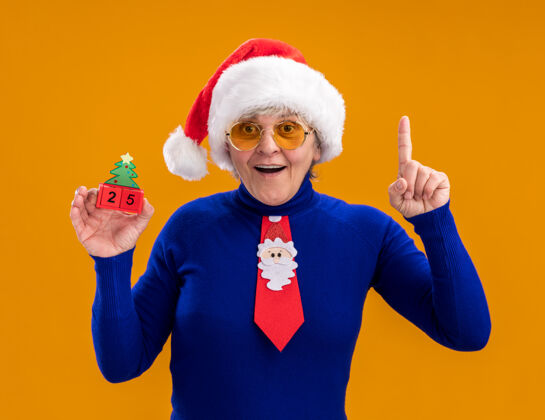 橘子戴着太阳眼镜 戴着圣诞帽 系着圣诞领带 手拿圣诞树饰物的惊讶的老妇人 指着橙色背景上孤立的复制空间圣诞老人老人装饰品