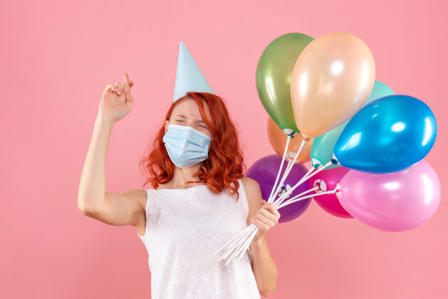 气球正面图是粉红色墙上戴着消毒面具手持彩色气球的年轻女子生日年轻女性快乐