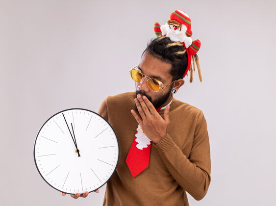 搞笑一个非洲裔美国人 穿着棕色毛衣 头上戴着圣诞老人的戒指 打着有趣的红色领带 手里拿着时钟 站在白色的背景上惊奇地看着它站着时钟非洲